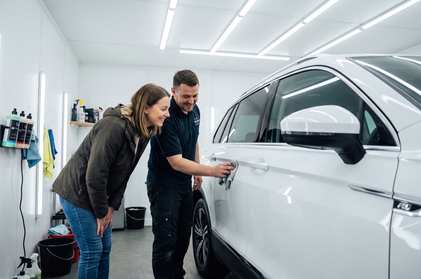 A detailer and a customer look at the door of a white SUV in a bright LED-lit detailing booth