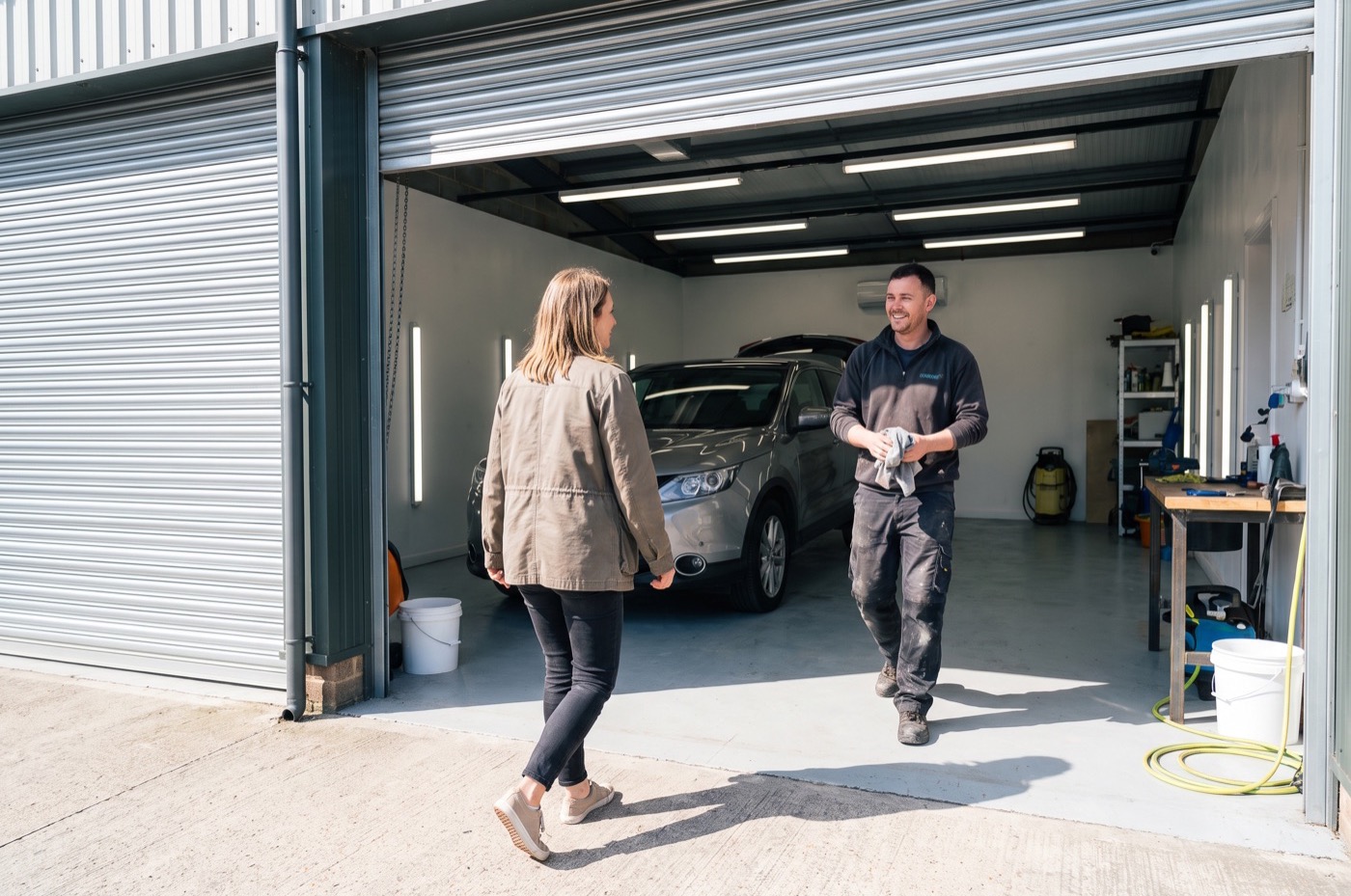 A customer walks into a detailing unit through the open roller shutter as the detailer wipes his hands and comes to greet her