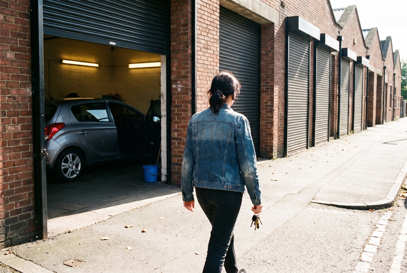 A woman with car keys in her hand walks along a sunny pavement past a row of brick units towards an open shutter with a hatchback inside