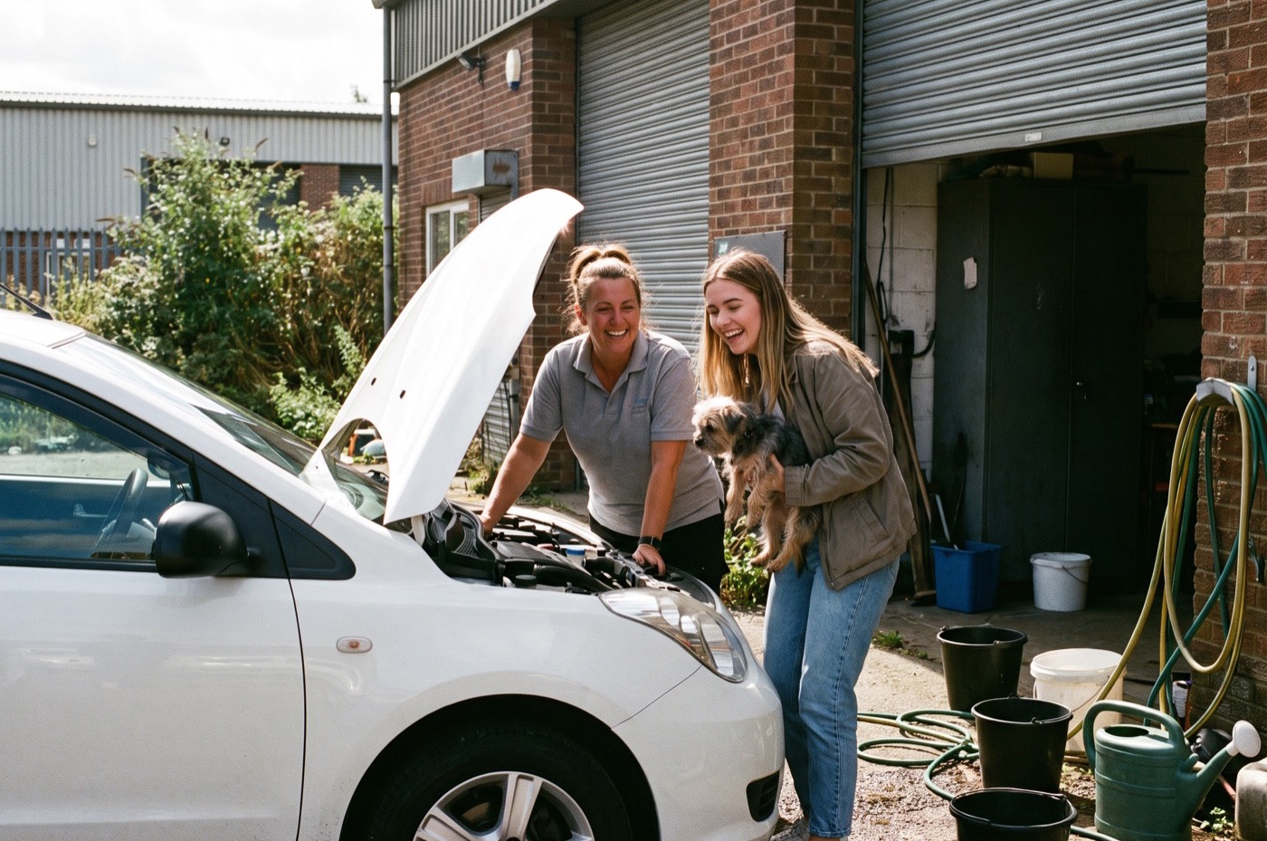 A female detailer and a young customer look at the bonnet of a small white hatchback parked outside a unit in the sun