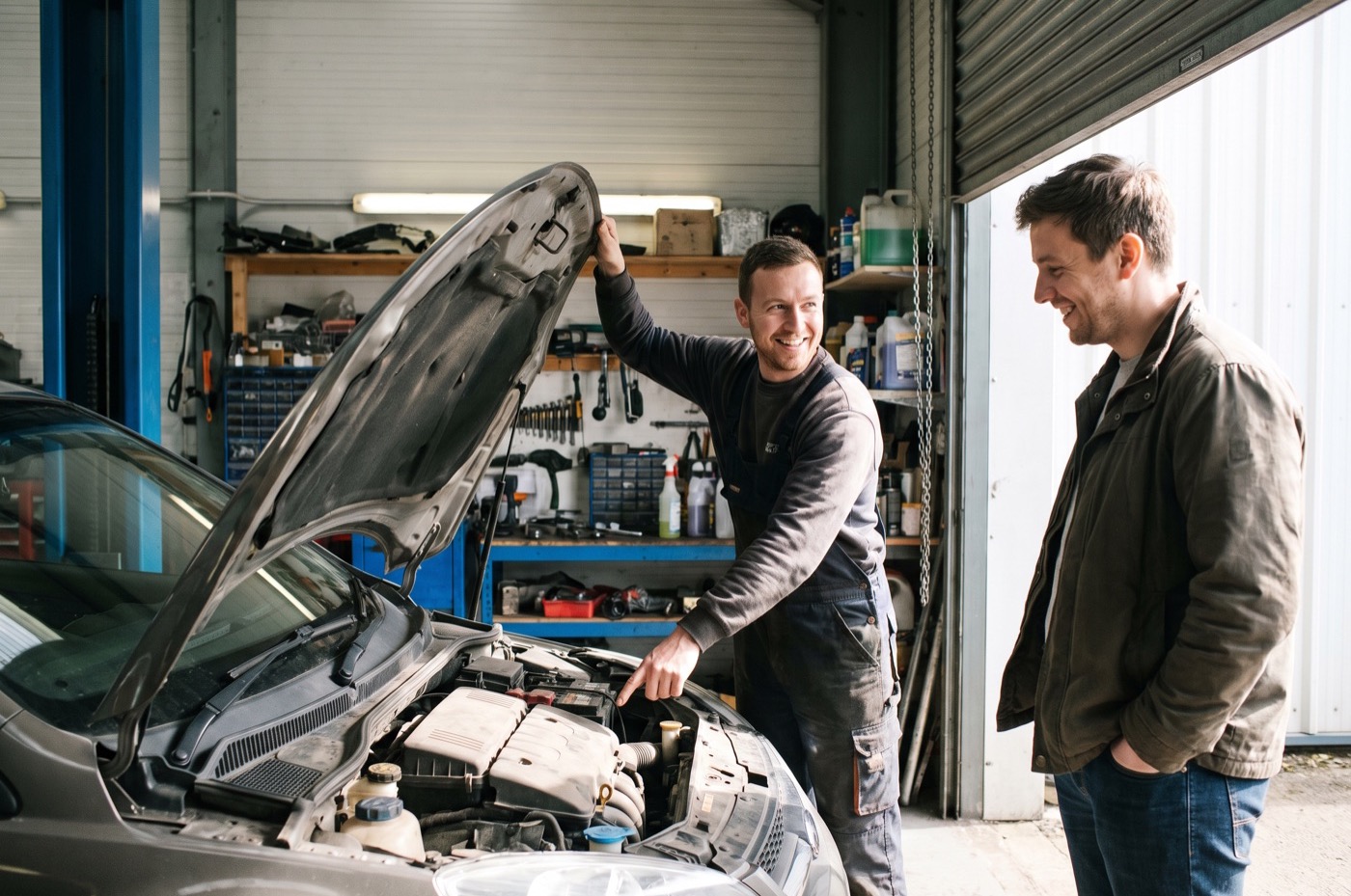 A detailer holds up the bonnet of a hatchback and points into the engine bay while the customer looks in