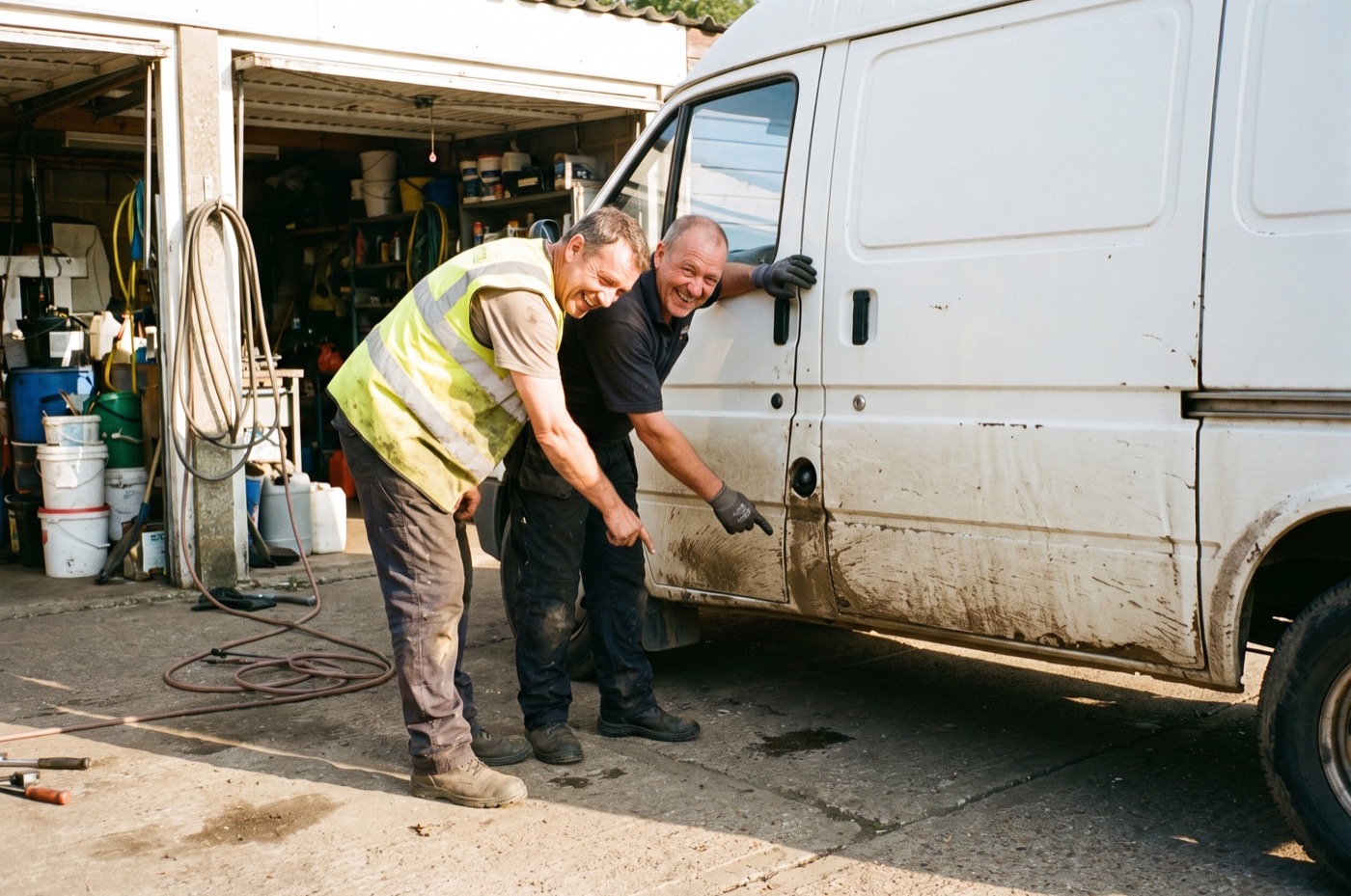 A tradesman in a hi-vis vest and a detailer laugh as they look at the mud on the sills of his white work van