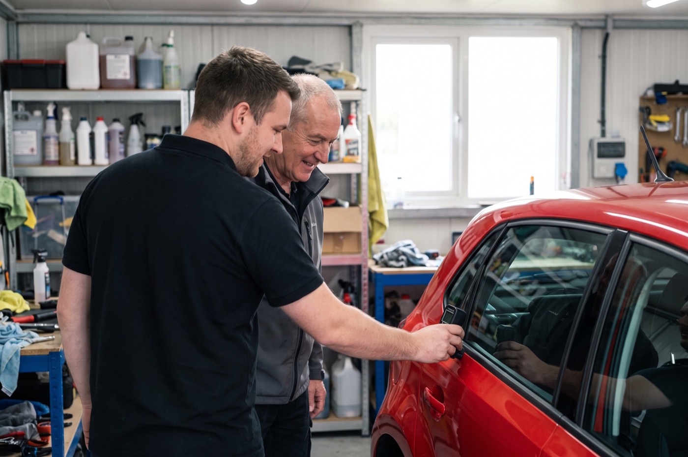 A detailer holds a paint-depth gauge against the door of a red hatchback while an older customer watches beside him