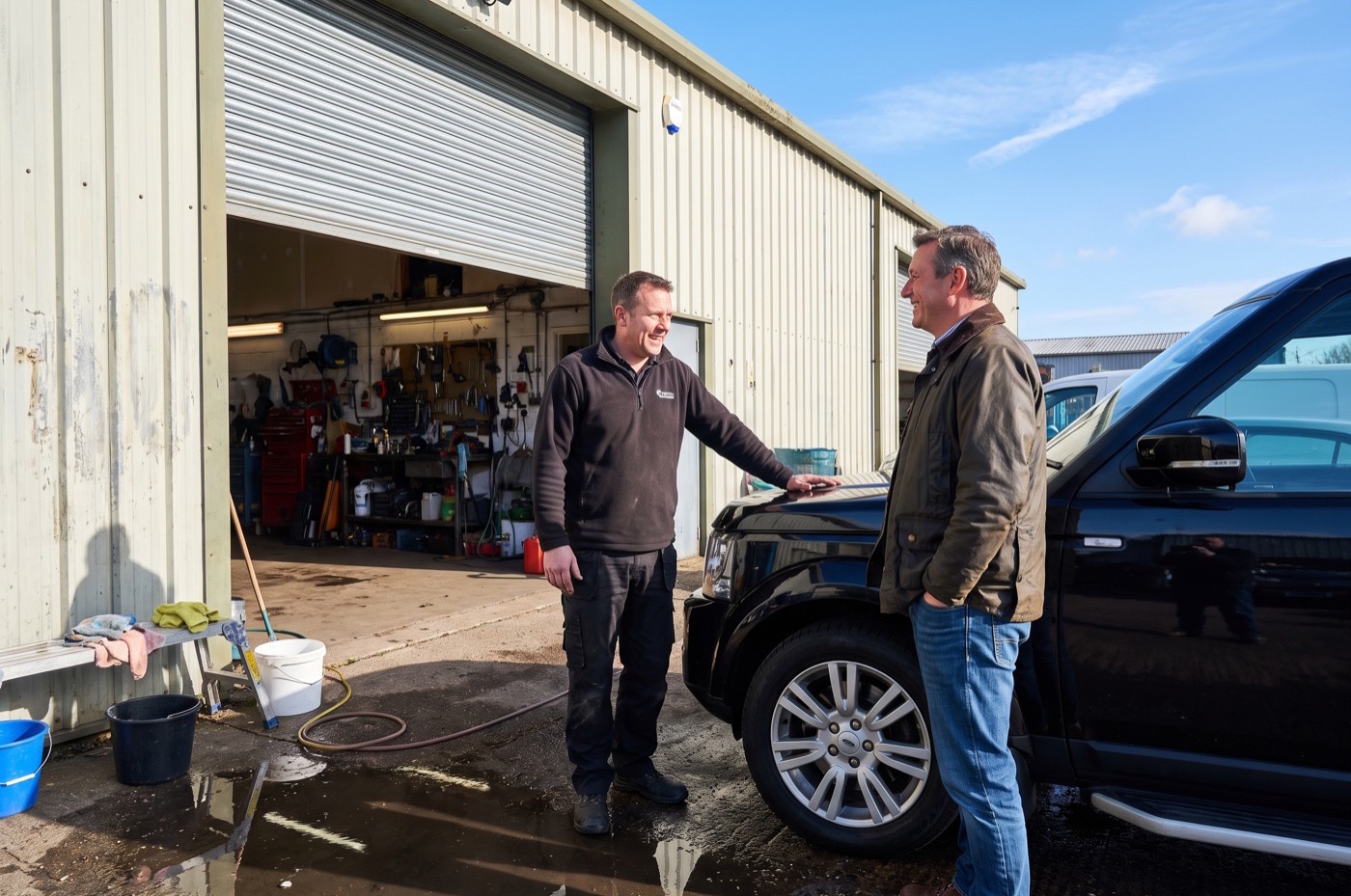 Two detailers and a customer talk beside a large black SUV outside an industrial unit on a bright morning