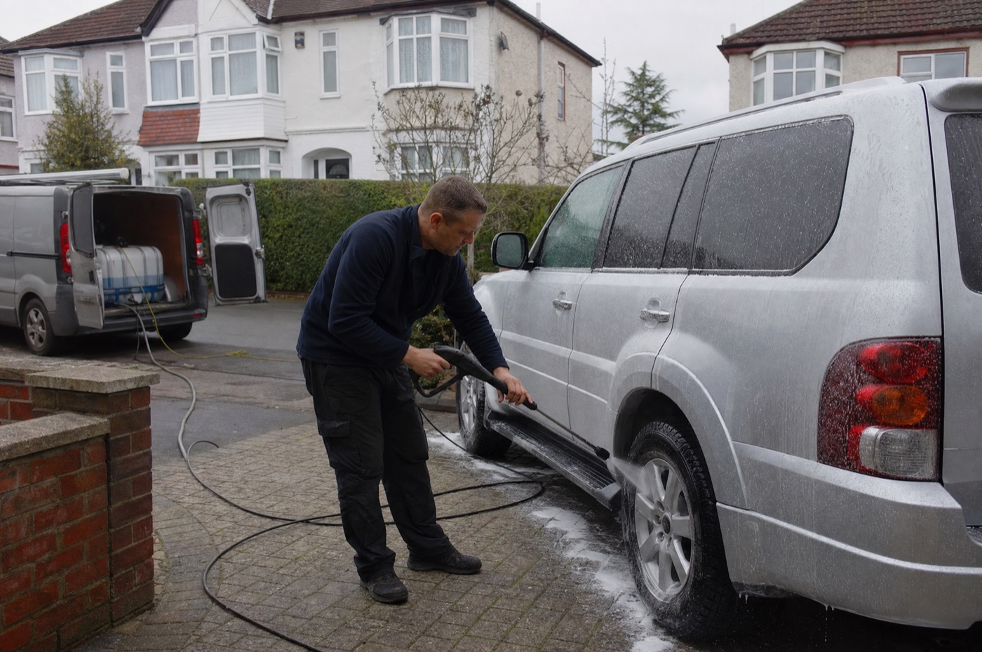 A mobile valeter rinsing the foam off a silver 4x4 on a suburban driveway, his van parked behind him